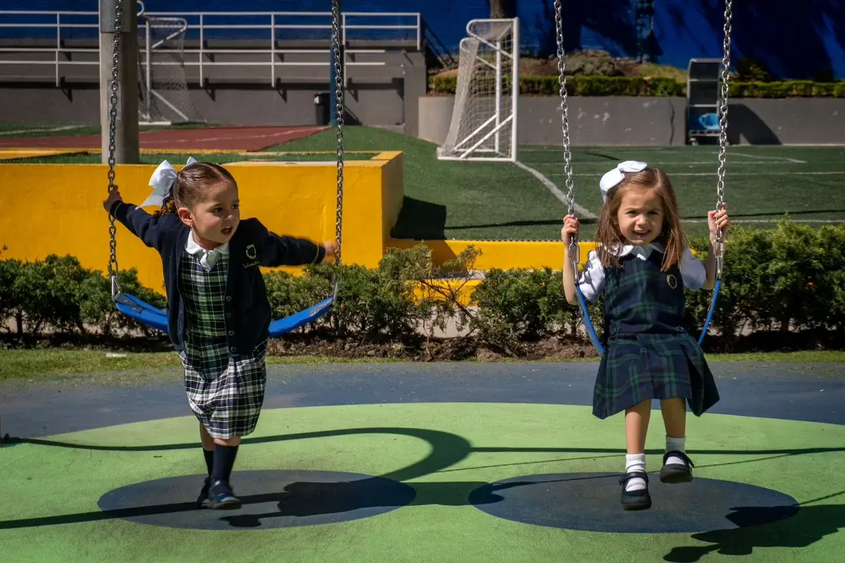 Dos estudiantes en uniforme escolar juegan en los columpios del área recreativa del colegio en un día soleado.