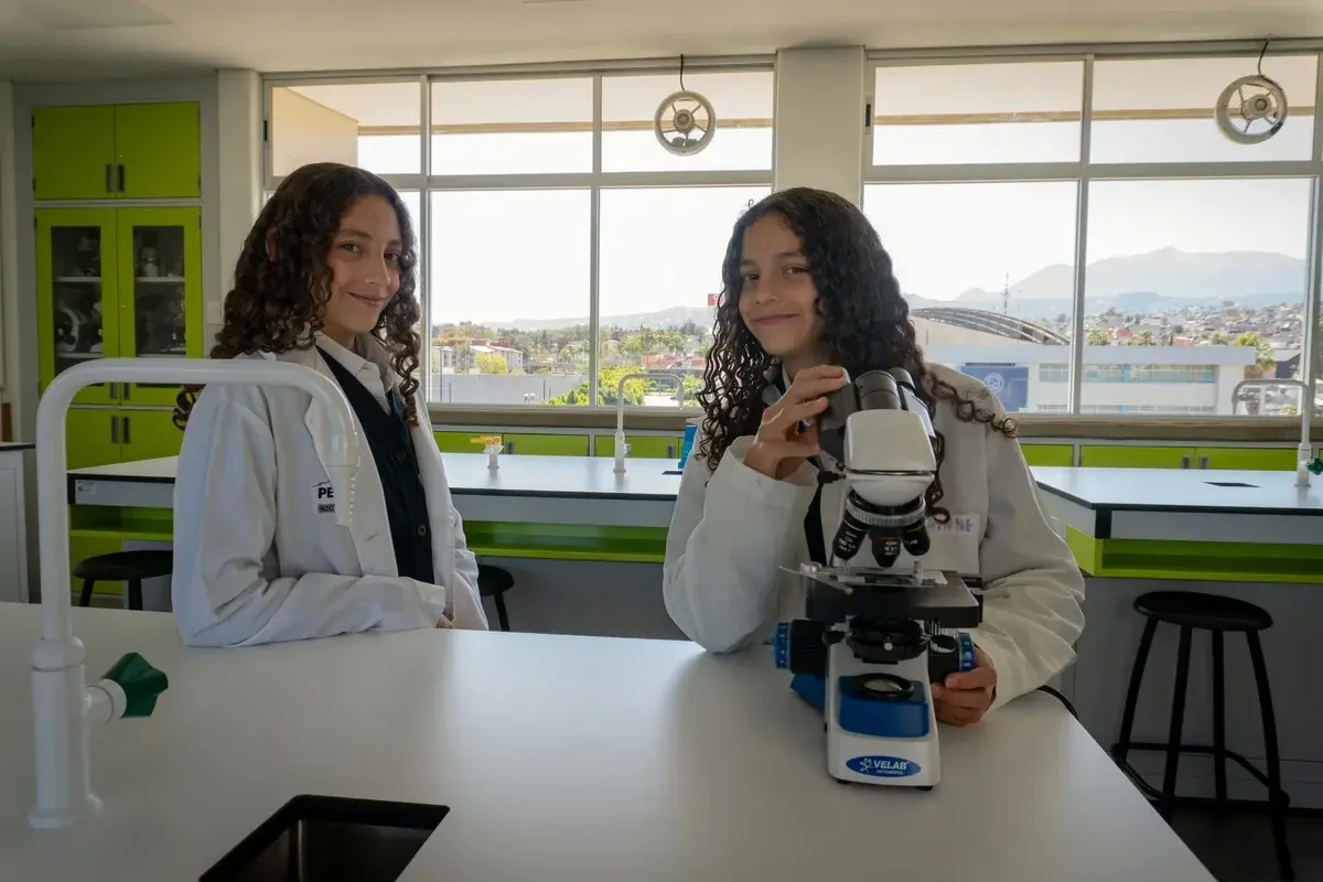 Dos estudiantes en uniforme observan un microscopio en el laboratorio con vista panorámica de la ciudad.